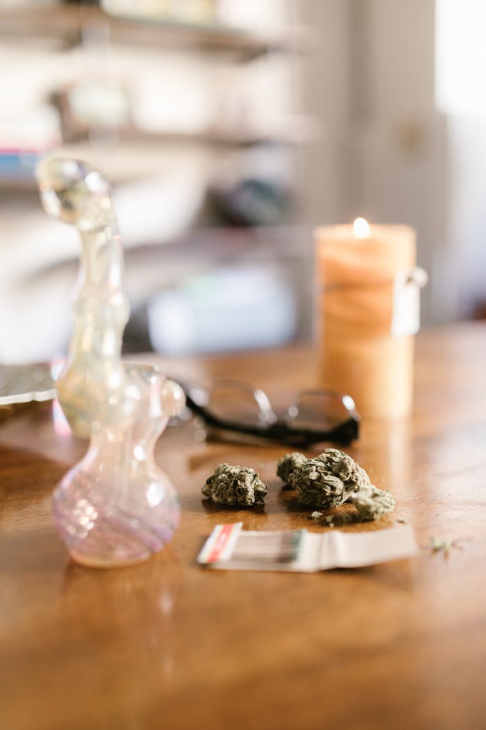 why-choose-us Cannabis flowers on a wooden table with a glass pipe and candle in a home setting.