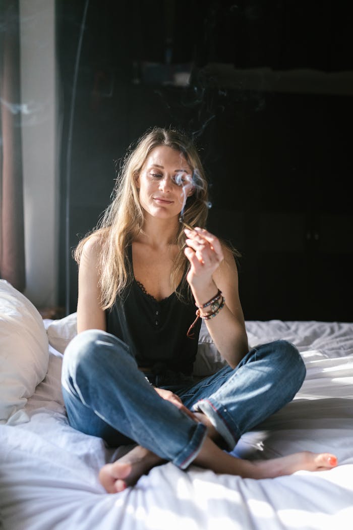 A young woman enjoying cannabis relaxation while sitting on a bed indoors.